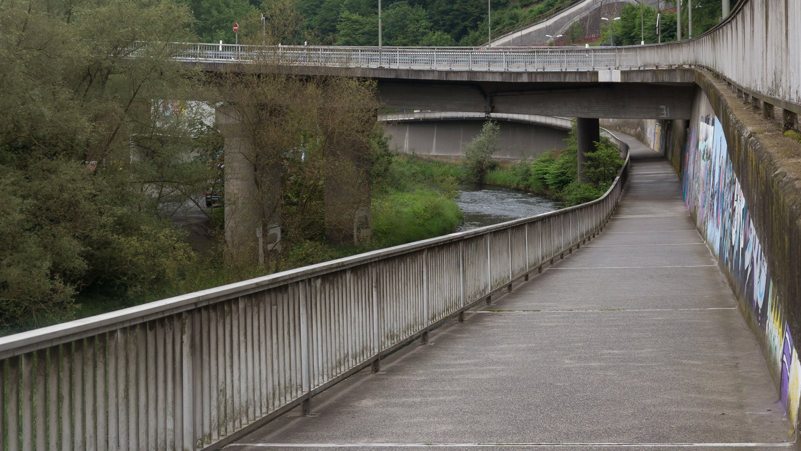 Wide cycle path curving along a river beneath road viaducts
